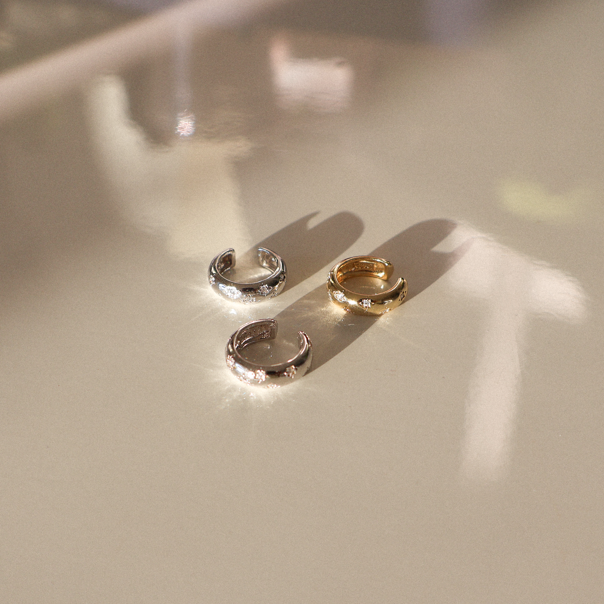 Three small crystal ear cuffs in silver, gold, and rose gold finishes arranged on a light surface with soft natural shadows.