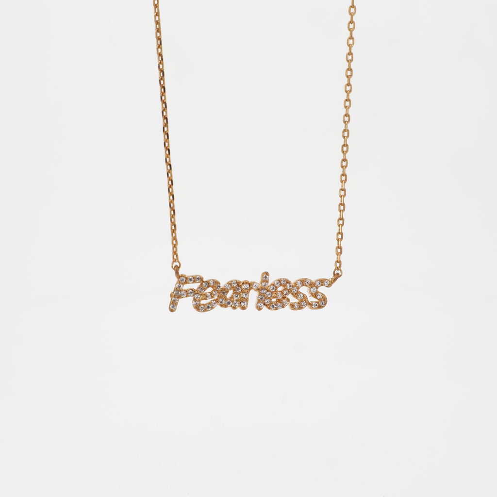 A gold-plated brass chain pendant necklace with the word 'FEARLESS' in bold script, featuring sparkling crystals.
